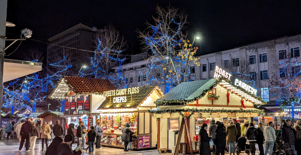Crowded outdoor Christmas market at night with wooden food stalls, festive lights in trees and people walking between the huts.